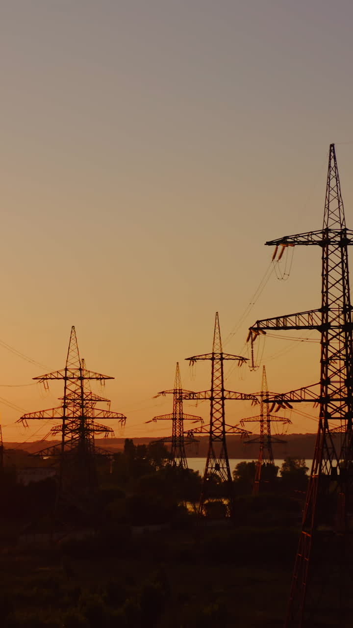 High voltage electrical lines at sunset. Dark silhouette of towers in the rays of the beautiful evening sun. Distribution and supply of electricity. Camera moves back. Vertical video