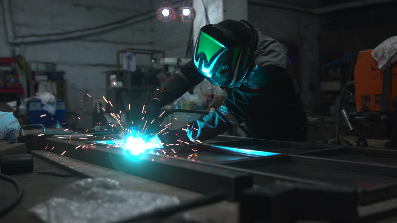 Adult worker in beanie and protective clothing bends over large steel panel while handheld tool sends bright sparks across dim workshop, creating intense industrial atmosphere with precision, heat