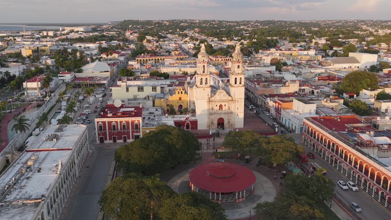 Aerial shot descending toward Campeche Cathedral and Plaza de la Independencia with colonial buildings and trees at sunset