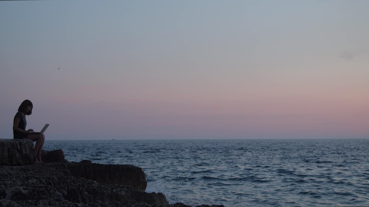 노트북이 바다 옆 바위에 앉아 있는 소녀, 그녀 위에 있는 새는 일몰, 넓은 촬영, 느린 동작으로 날아가고 있습니다.