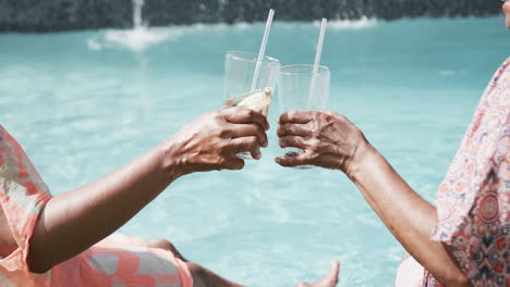 midsection of senior african american female friends sitting by pool making a toast, slow motion