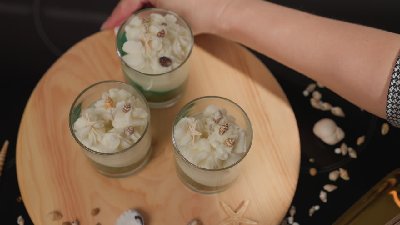 Female chandler carefully adjusts round wooden tray using both hands to position three glass jars styled like whipped desserts with shell toppings, surrounded by scattered decorative seashells