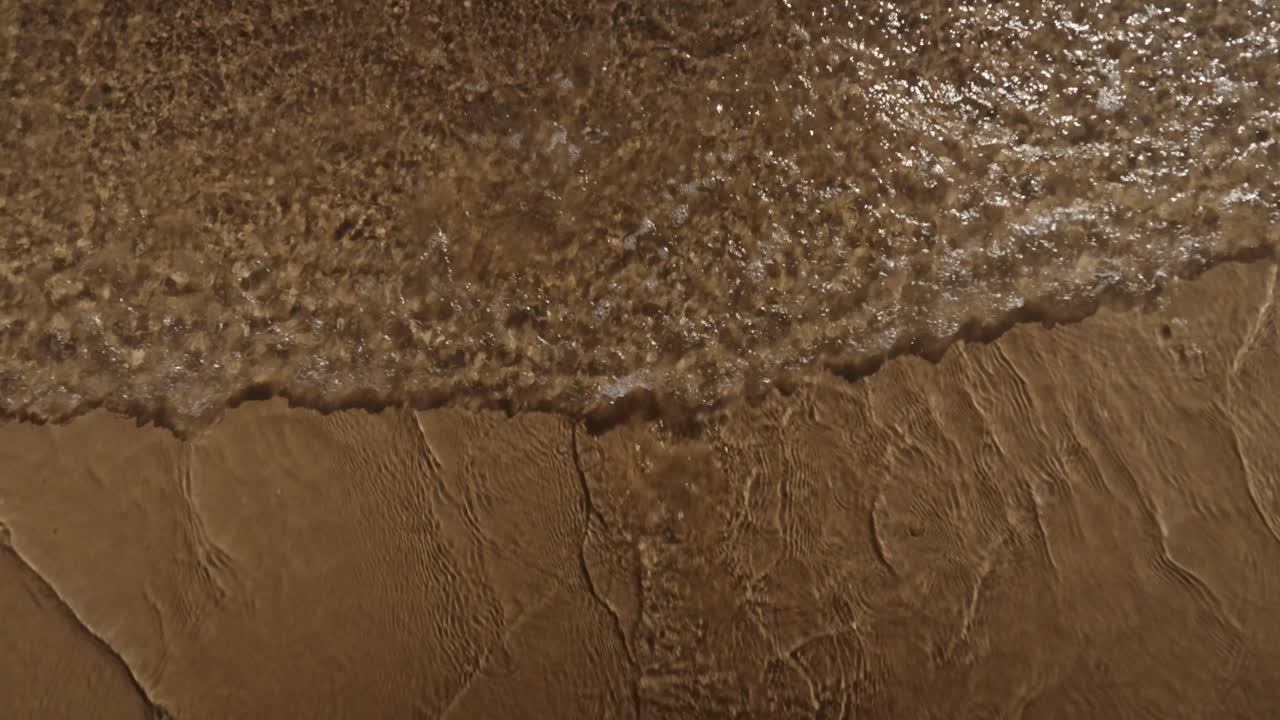 A gentle wave rolls onto rippled sand, capturing textures and light reflections along the Algarve shoreline in Portugal