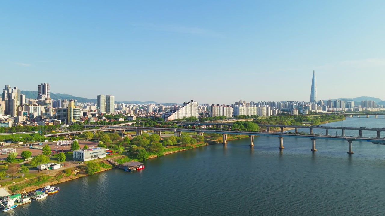 Sorvolo aereo lungo il fiume Han a Seul che passa i ponti Gwangjin e Cheonho, con l'iconica Lotte World Tower che si erge sopra lo skyline della città sotto un cielo diurno sereno.