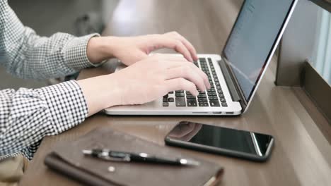 young asian business man using laptop computer in working space with smartphone and notebook on wooden desk. male hand typing on laptop keyboard. freelance lifestyle in digital age concept.