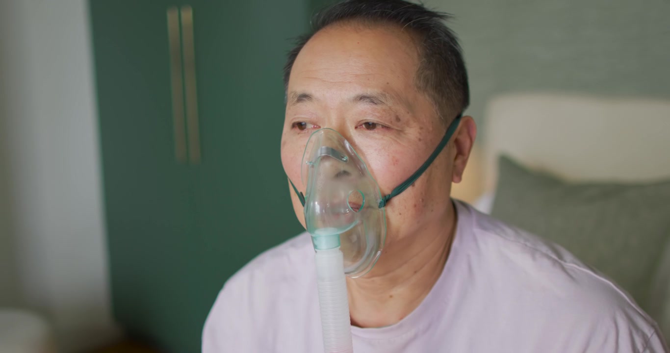 Anziano uomo asiatico che riceve ossigeno tramite maschera e tubi, respirando regolarmente a letto per supporto