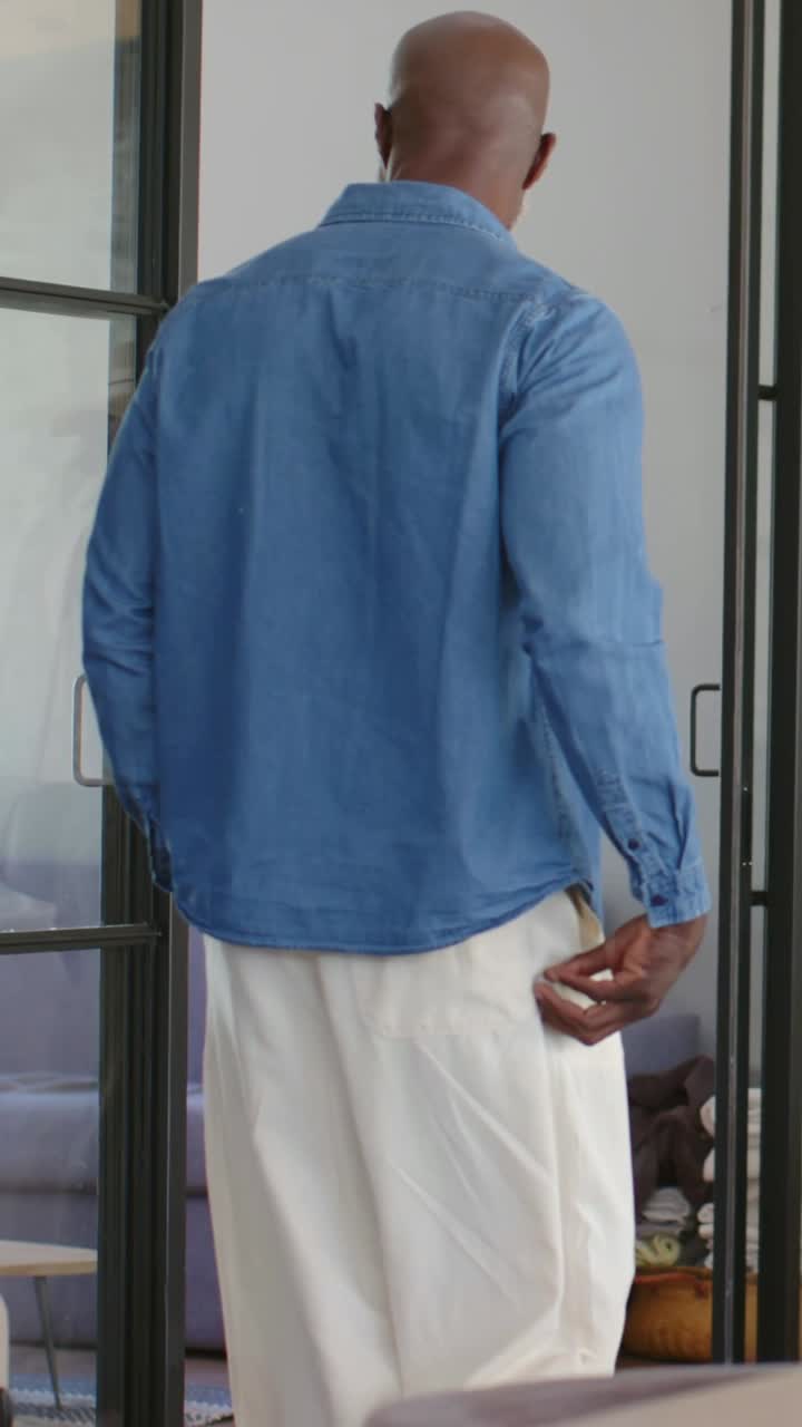 Video verticale: Uomo che cammina in camicia di jeans e pantaloni color cammello, si sposta verso una porta a vetri di casa, con una valigia.