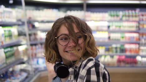 Supermercado:-Niña-Feliz-Bailando-Divertido-Entre-Estantes-En-El-Supermercado.-Chica-Rizada-Con-Camisa-A-Cuadros-En-Blanco-Y-Negro-Con-Auriculares-En-El-Cuello.-Camara-Lenta.-De-Cerca