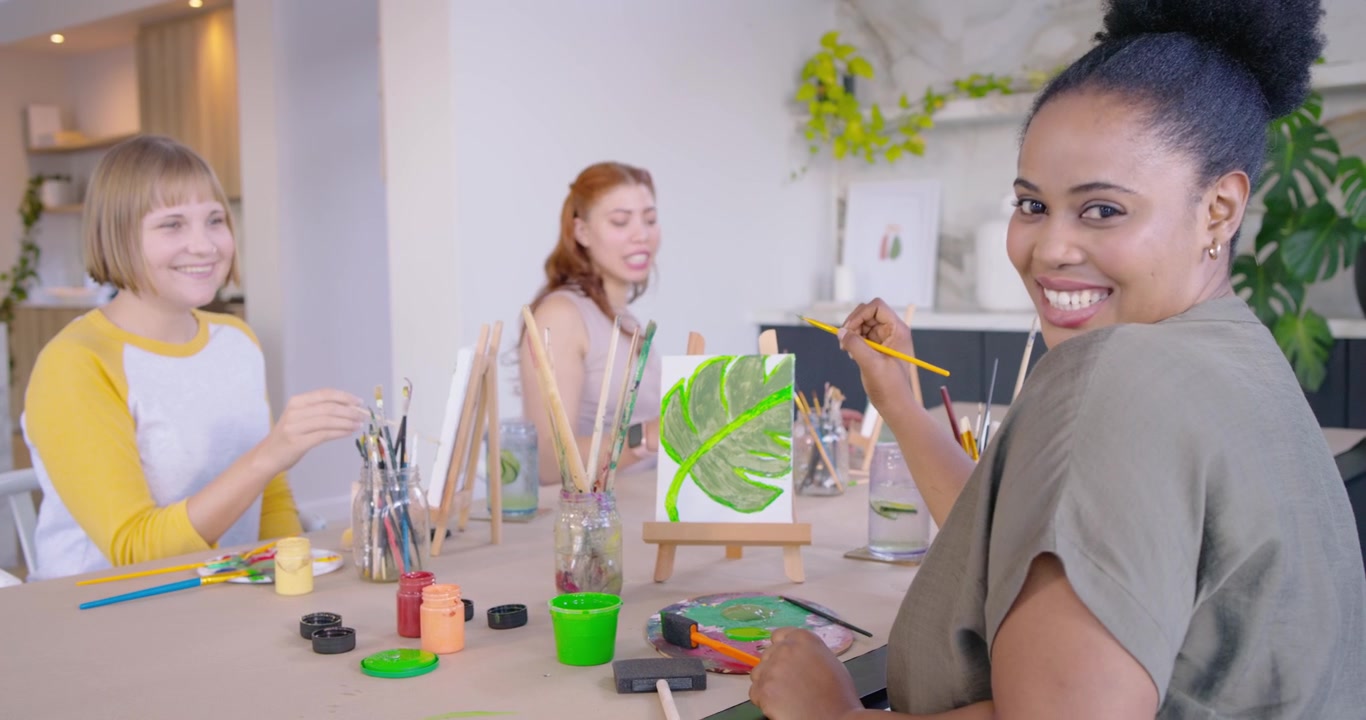 Mulheres diversas pintando tela em casa, a do centro falando, a mulher da direita, de blusa verde-oliva, refinando uma folha para a aula.