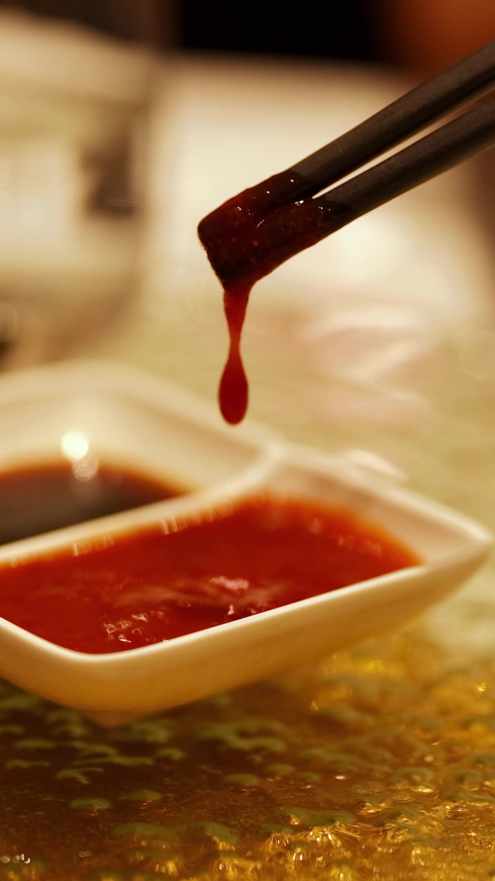 暖かな照明の中華料理店で、箸が皿の中の赤いソースをかき混ぜる瞬間を捉えた一枚。