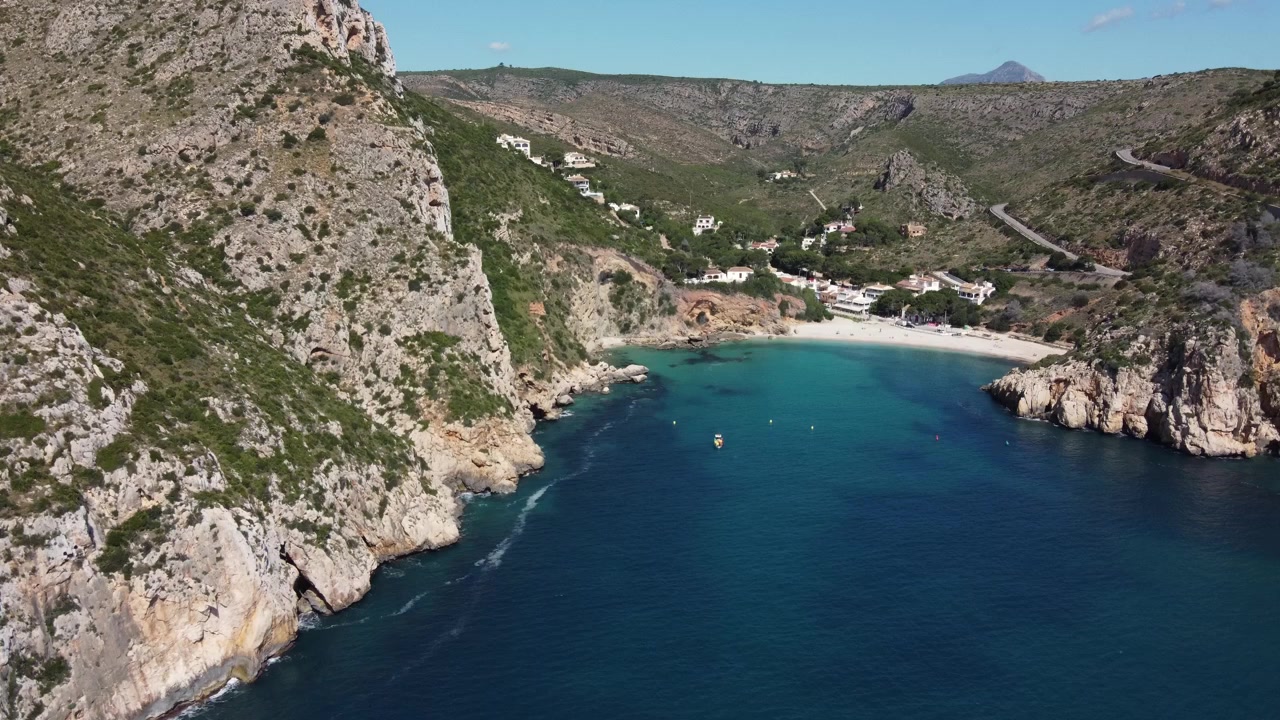 Malebný letecký vzlet dronu nad tyrkysovými vodami zátoky Granadella v Javea ve Španělsku, postupně odhalující svěží středomořské údolí a zvlněné kopce v pozadí.
