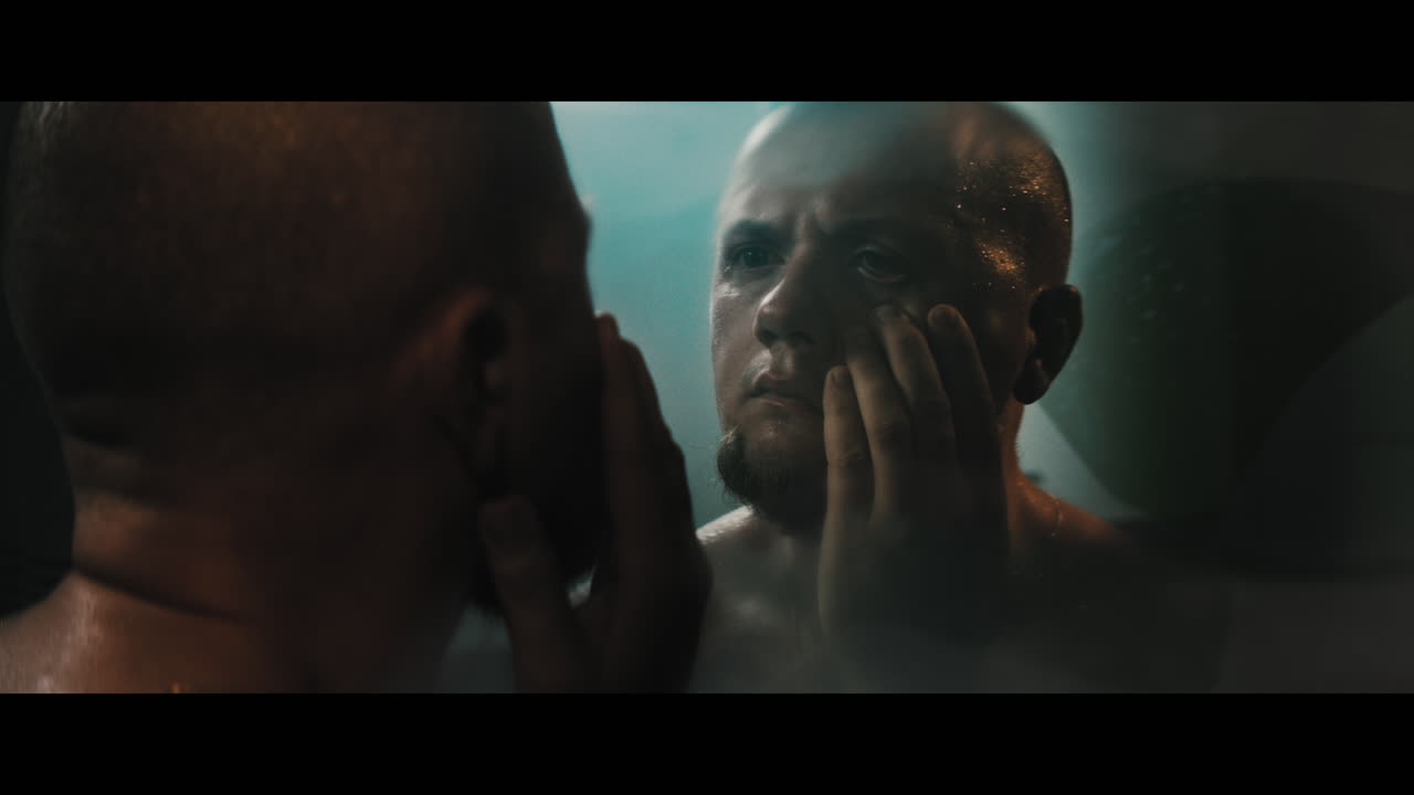 A bald dangerous looking man looks on his face in the bathroom mirror