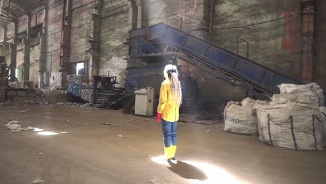 imágenes de una mujer con rastas con chaqueta amarilla y botas de pie en el medio del área vacía de la fábrica de reciclaje de residuos en los rayos de luz natural de cieling