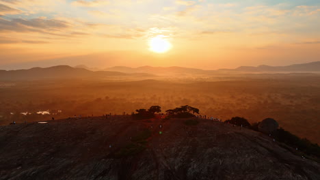 lots of people stand on the high rounded rock with little vegetation. hazy scenery with mountain silhouettes at backdrop. tourists watch the beautiful sunset over sri lanka, south asia.