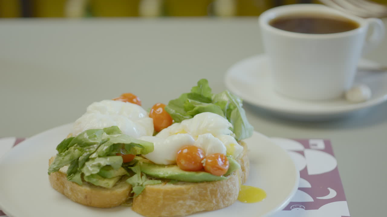 Close-up of a plate with rustic bread toast topped with avocado, lettuce, cherry tomatoes and poached eggs, next to a cup of hot coffee
