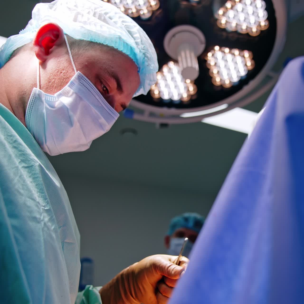 Medico uomo di mezza età con mascherina e cappellino. Ritratto da un'angolazione bassa di un chirurgo intento al lavoro.
