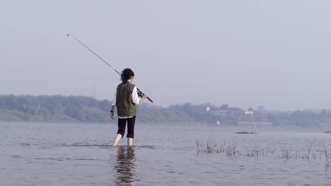 Seorang wanita Asia Selatan berjalan melewati air danau yang dangkal sambil memegang pancing dan melihat sekeliling, menikmati ketenangan alam pedesaan dan cahaya siang hari