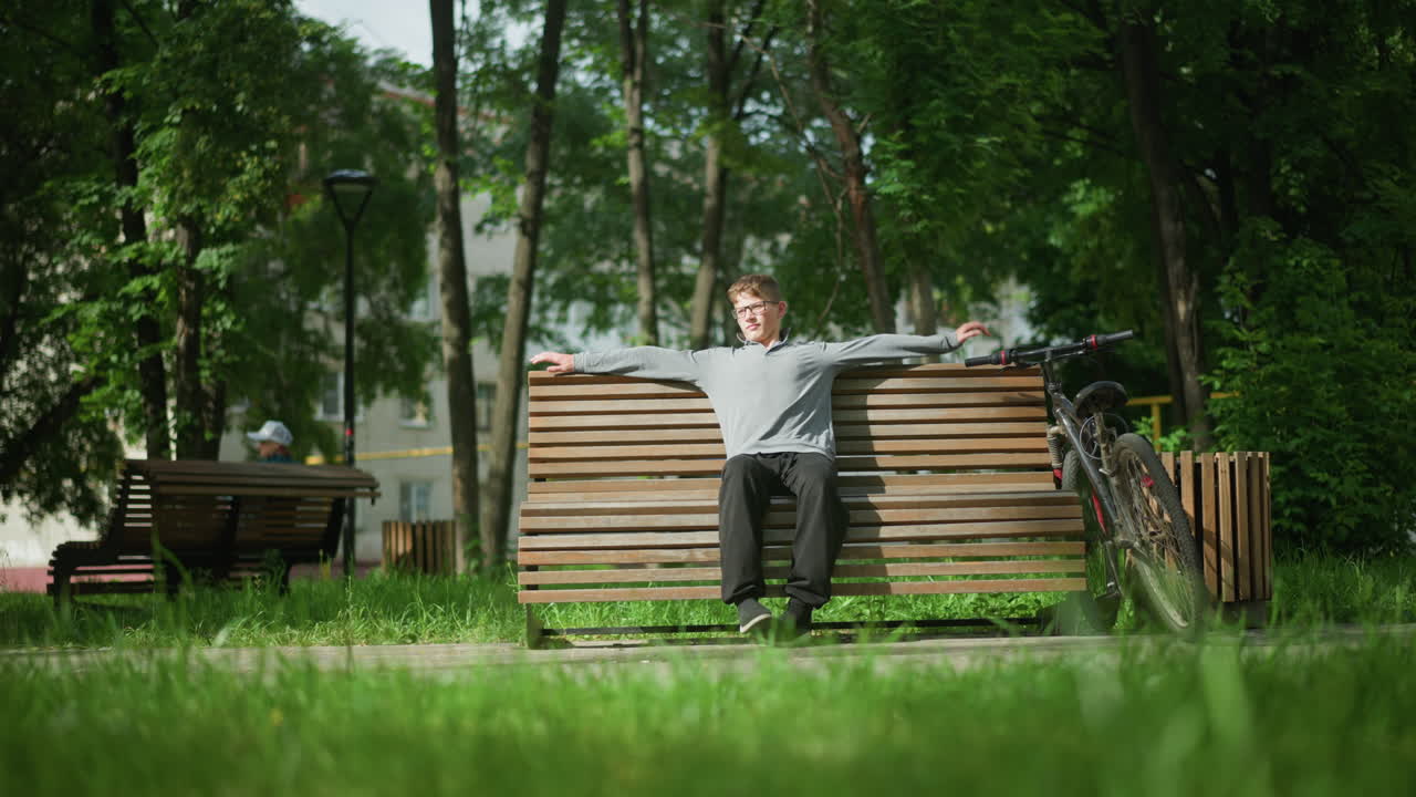 하 소년은 밖에서 나무 벤치에 앉기 전에 뒤로 돌아보고, 벤치 위에 어, 왼쪽 다리에 오른쪽 발을 놓고, 그의 에 주차 된 자전거, 다른 사람이 멀리 벤치에서 앉아 있습니다.