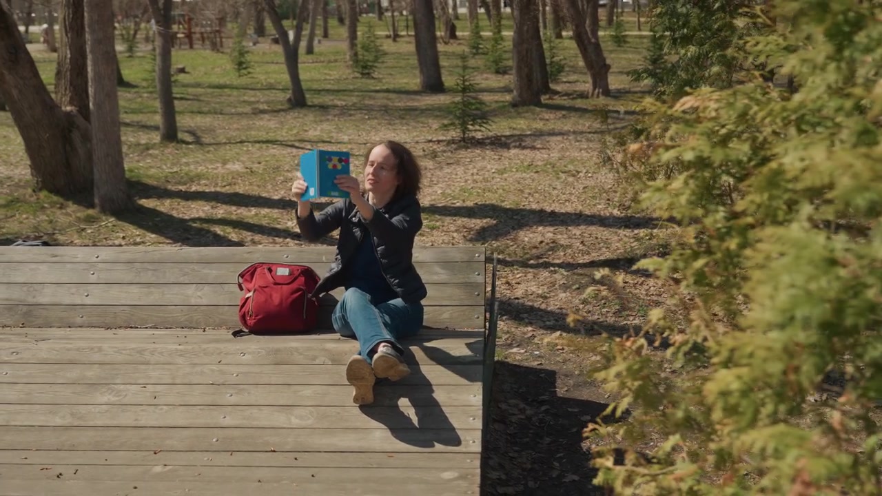 Donna legge un libro su una piattaforma di legno in un parco, tenendo un quaderno blu con copertina colorata e uno zaino rosso accanto a lei. Mentre la donna è seduta a gambe incrociate, la luce del sole proietta ombre da alberi e foglie.