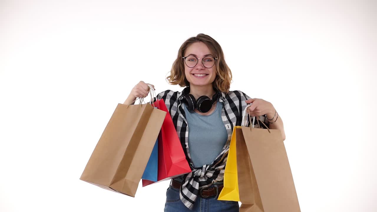 안경과 헤드폰을 목에 고 계절 판매에서 다채로운 쇼핑 가방을 들고있는 유쾌한 아름다운 유럽 젊은 여성은 색 배경에 고립 된 은 손으로 행복하게 보여줍니다.