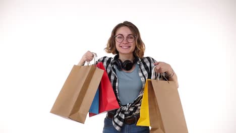 alegre hermosa joven europea con gafas y auriculares en el cuello sosteniendo bolsas de compras de colores en la venta de la temporada, felizmente mostrarlos en las manos extendidas aislados en fondo blanco