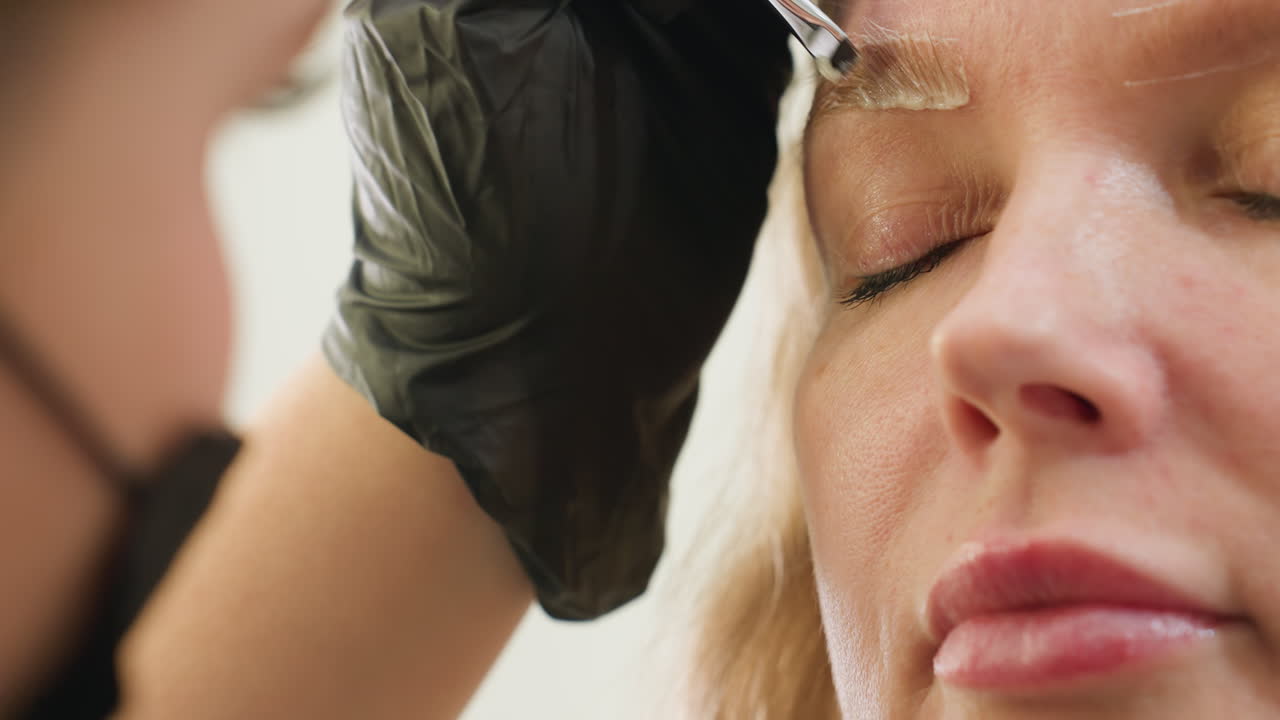 Close-up of client with closed eyes while technician gently applies mixed substance on eyebrow using small angled brush, technician slightly blurred in foreground wearing black gloves and face mask