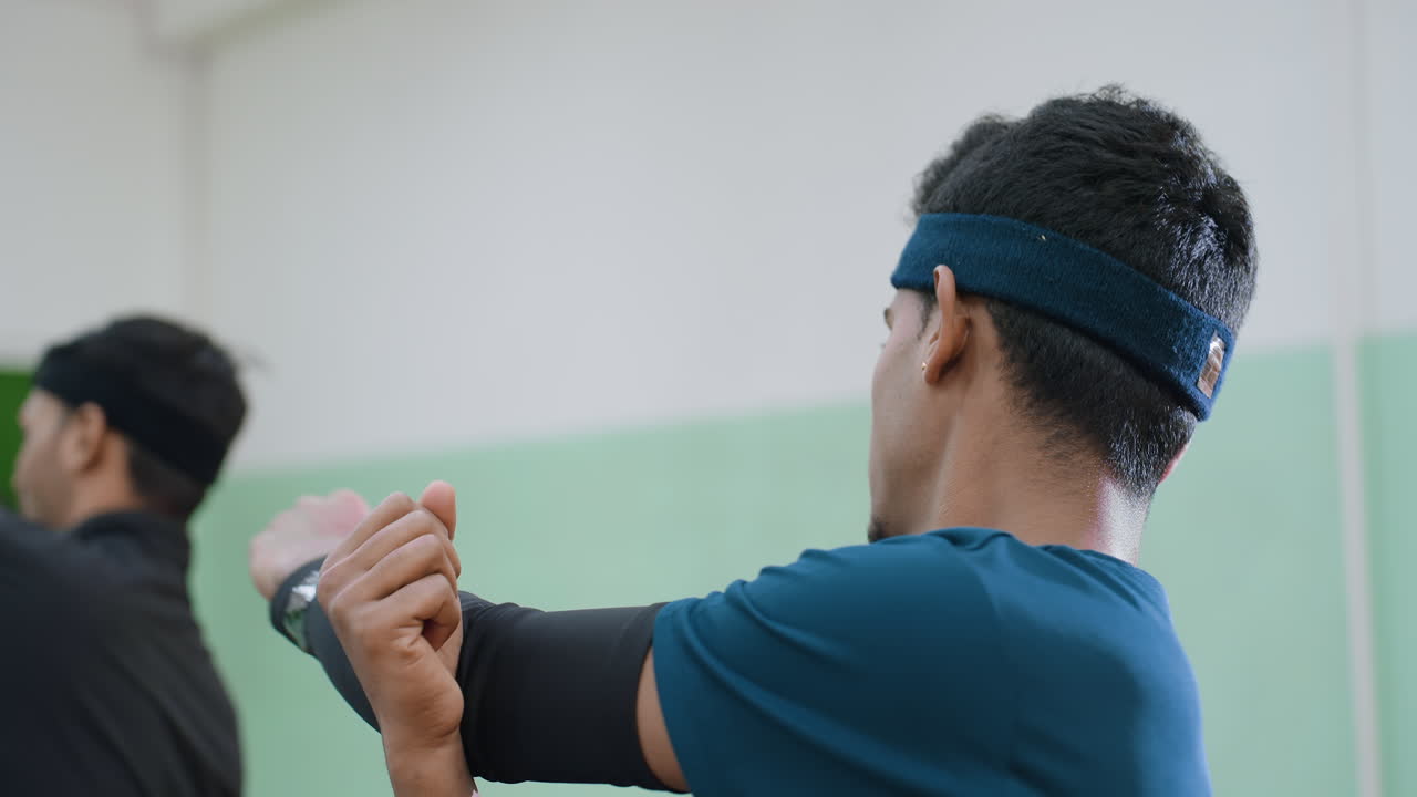 Close up of two athletes wearing headbands engaged in arm stretching during indoor training session, focusing on flexibility and preparation before sports performance in gym hall