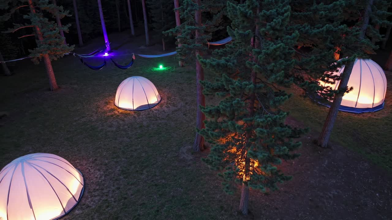 Several illuminated glamping domes and hammocks nestled in a pine forest at dusk, creating a serene and inviting atmosphere for a unique outdoor experience