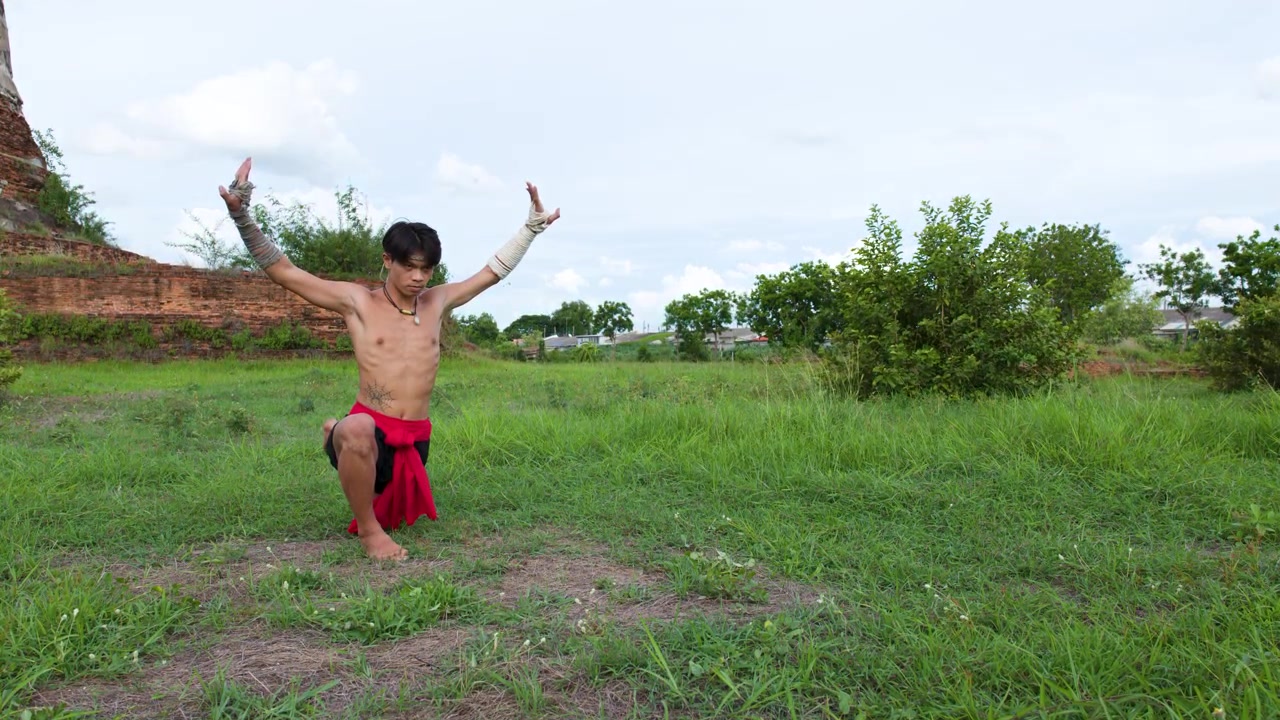 Shirtless fighter in red sarong performs ancient martial arts ritual outdoors near historic ruins.