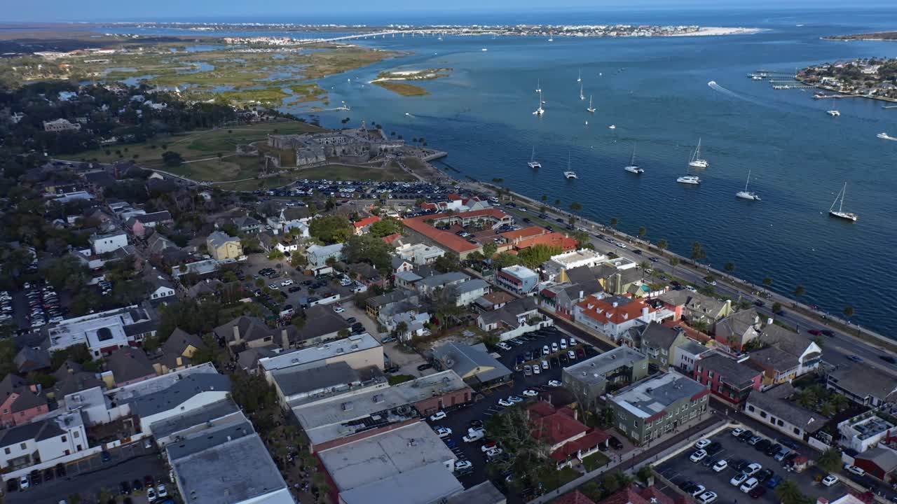 Cittadina costiera con un porticciolo di barche a vela ancorate vicino a una strada sul lungomare, con edifici residenziali e commerciali adiacenti a zone paludose e acque aperte sotto un cielo sereno