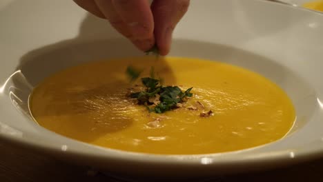 a white male hand is sprinkling herbs on a yellow soup in a white plate in slow motion