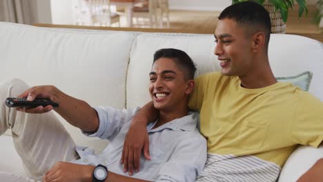 Smiling-mixed-race-gay-male-couple-sitting-on-sofa-watching-tv-one-using-remote