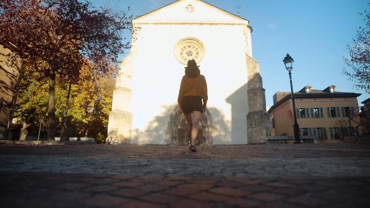 Brunette woman wearing a hat, walking towards a church in autumn sunset lights