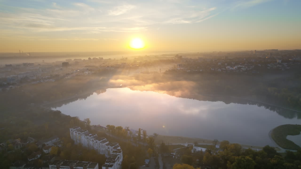Aerial drone view of Chisinau at sunrise, Moldova. View of Valea Morilor Park with lake and yellowed forest, multiple buildings around, fog in the air