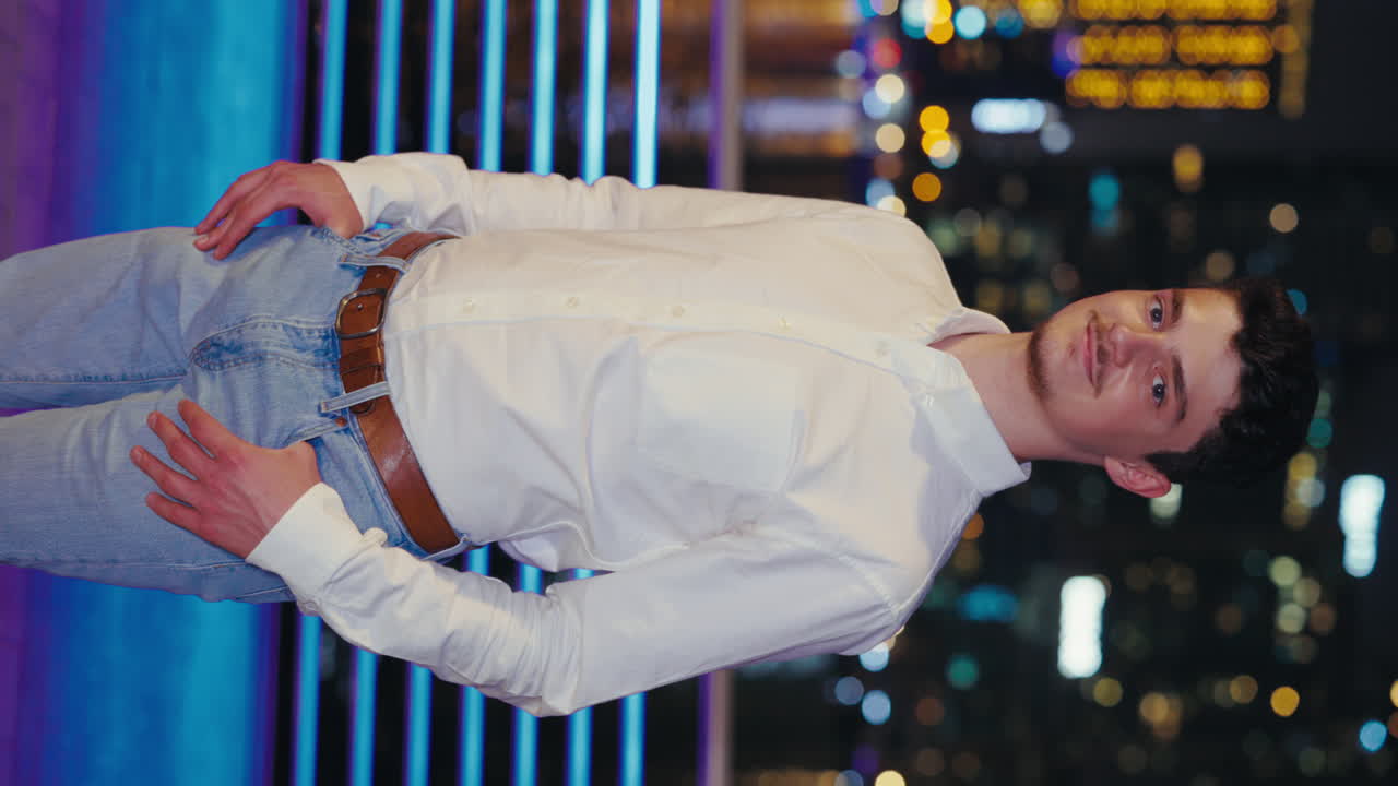 Man in white shirt and jeans posing in front of city lights