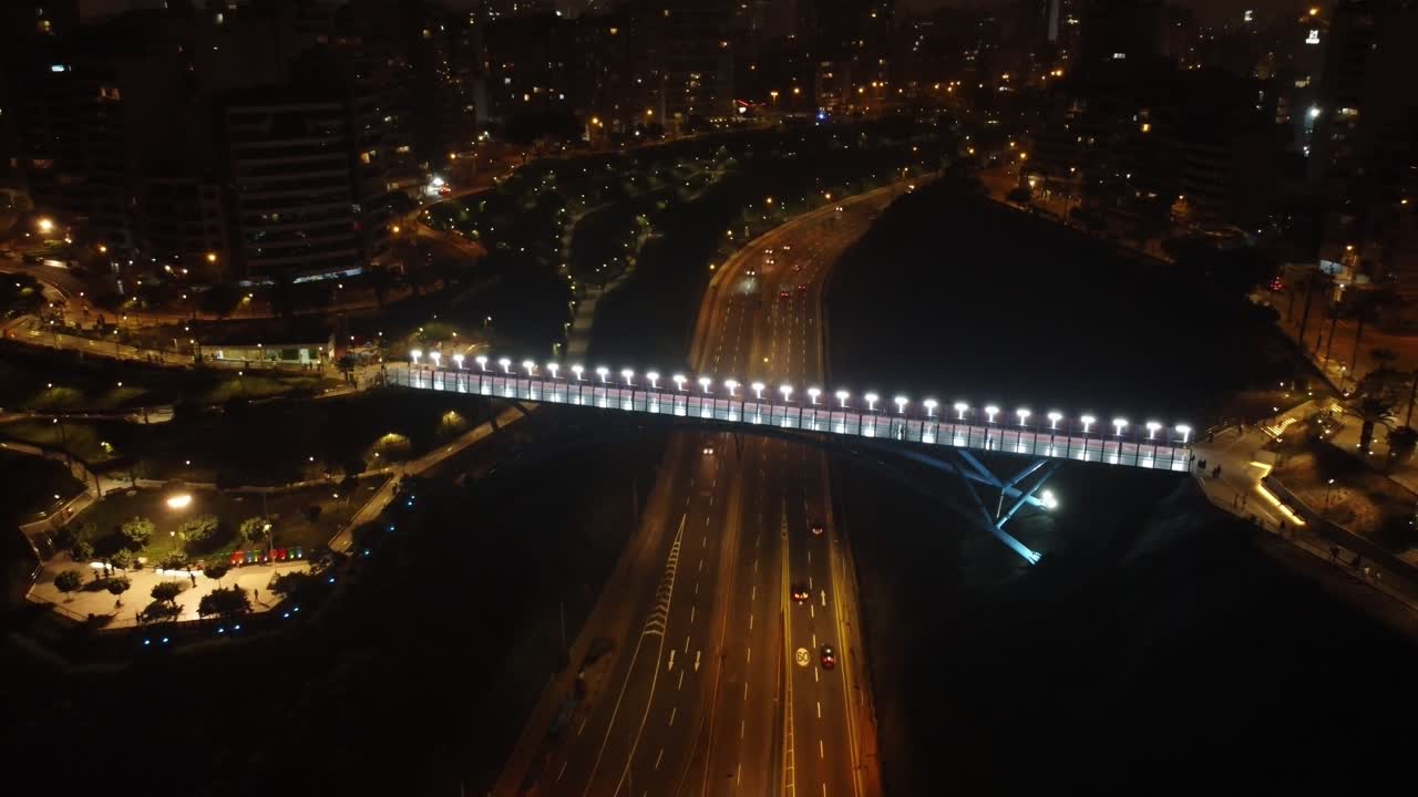 Aerial night time shot of a bridge above a street called "Bajada Armendariz". The drone flies backwards and up, away from the brightly lit bridge. Location is in Lima, Peru