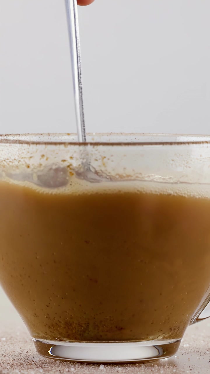 Primo piano di un cucchiaio di metallo che mescola caffè caldo con schiuma in una tazza di vetro trasparente.