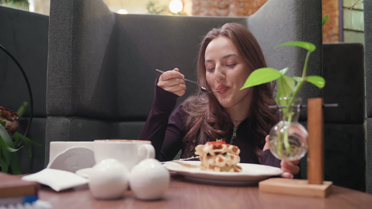 Lady enjoys waffles with gentle smile while seated at cozy booth, holding fork near mouth. Decorative plant in glass vase and white round ornaments add charm to tranquil indoor dining moment