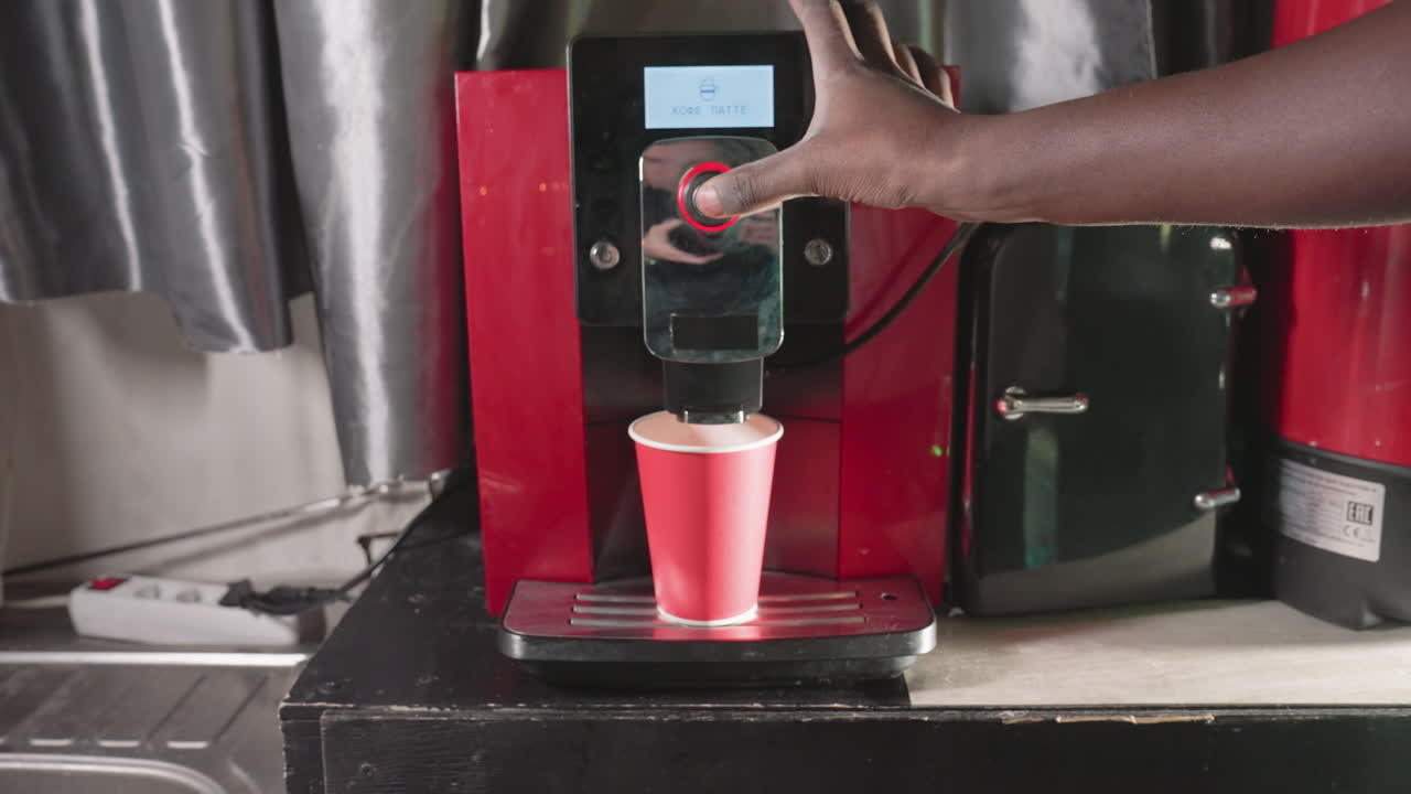 Close-up shot of a hand placing a red paper cup under an espresso machine's nozzle. The machine is set up to dispense coffee, with its sleek design and digital interface visible in the background