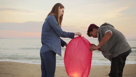 Junges-Multiethnisches-Paar-Zündet-Vor-Dem-Start-Eine-Blitzrote-Papierlaterne-An.-Romantisches-Date-Am-Strand.-Attraktive-Frau-Hält