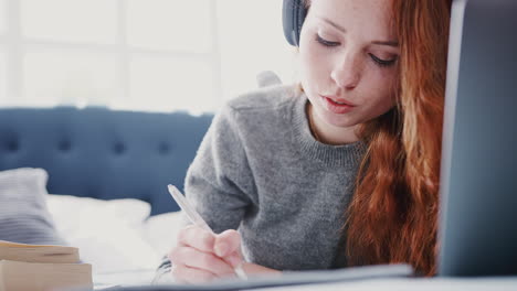 Female-College-Student-Wearing-Headphones-Works-On-Bed-In-Shared-House-With-Laptop-And-Mobile-Phone