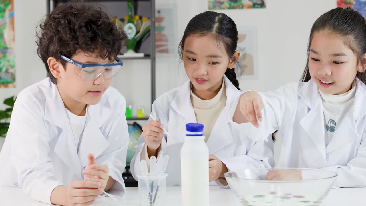 3人のアジア人学生が実験衣と安全ゴーグルを着用し、協力して化学実験を行っている。