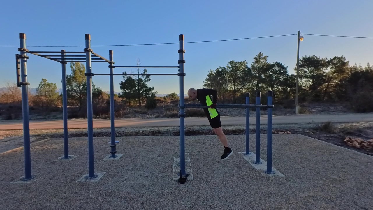 Vista lateral de un hombre atlético haciendo fondos de tríceps en barras paralelas en una zona de fitness al aire libre. Luz solar matutina con árboles de fondo. Concepto de entrenamiento con peso corporal, fuerza, al aire libre.