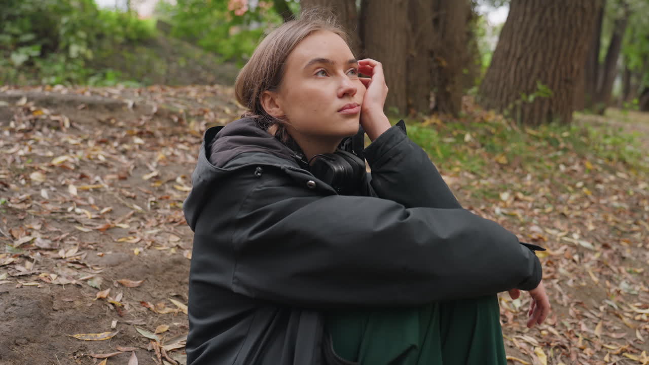young white woman sitting forest in oversized black jacket and green trousers, wearing headphones, gazing upward among oak trees and fallen leaves, quiet reflective mood, soft overcast light, candid