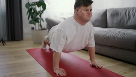 Hombre-Discapacitado-Enfocado-Con-Síndrome-De-Down-Haciendo-Flexiones-En-La-Alfombra-En-La-Sala-De-Estar