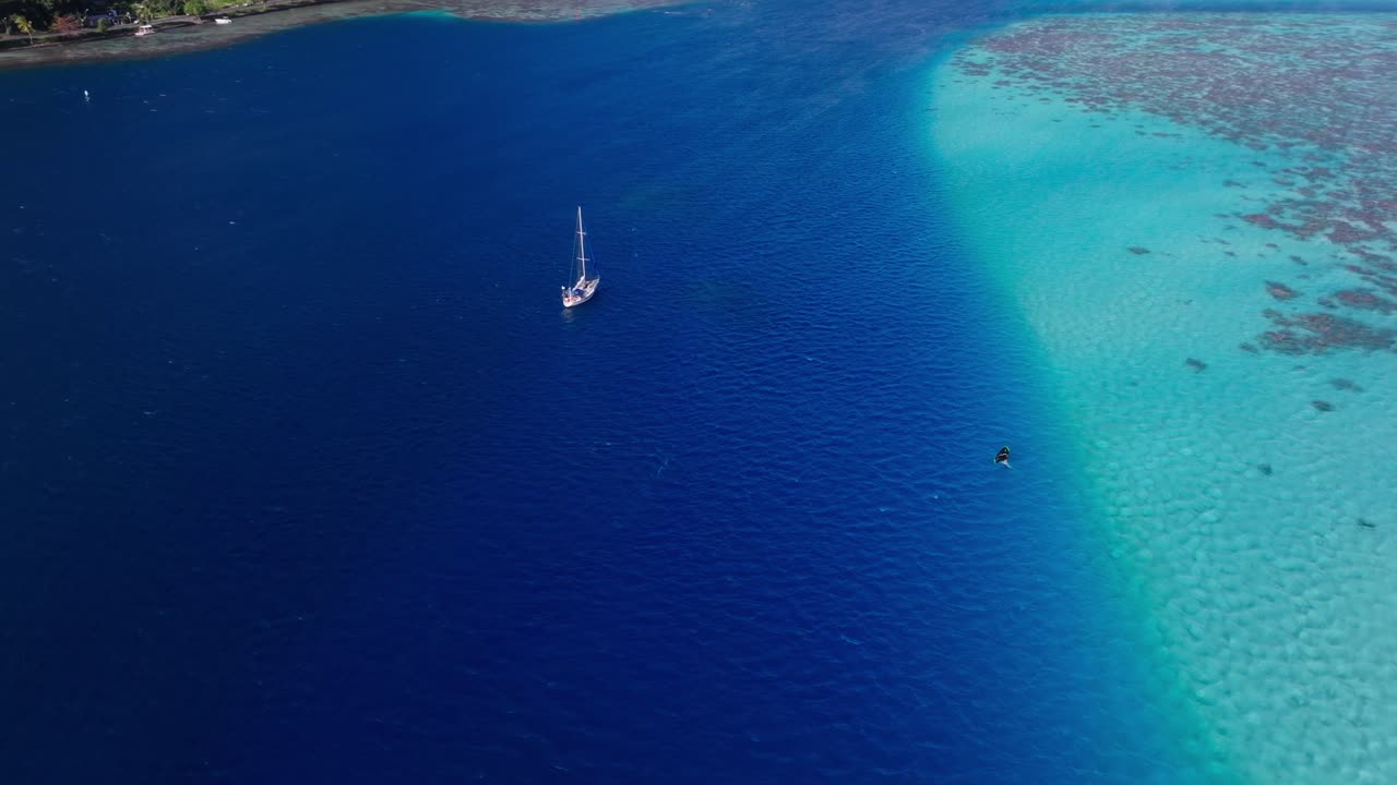 A windsurfer enjoys a sunny day on the waters while a sailboat is passing by. They are surrounded by coral reefs of Tahiti, French Polynesia