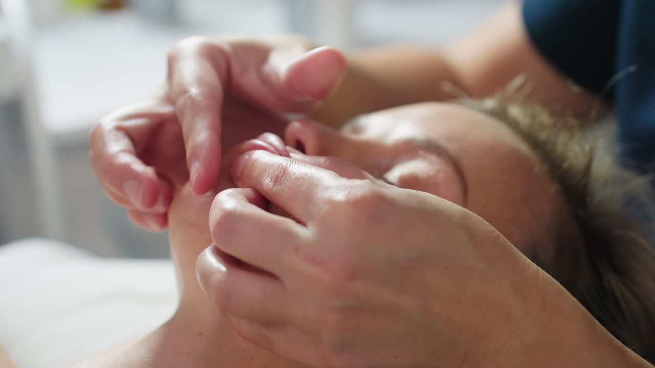 Close up of relaxed client lying with eyes closed in spa treatment as beauty expert gently massages lips and cheeks with nourishing cream using soft strokes to rejuvenate and hydrate skin