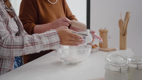 Primer-Plano-De-Las-Manos-De-La-Abuela-Poniendo-Harina-En-Un-Colador-Mientras-La-Nieta-Tamiza-El-Ingrediente