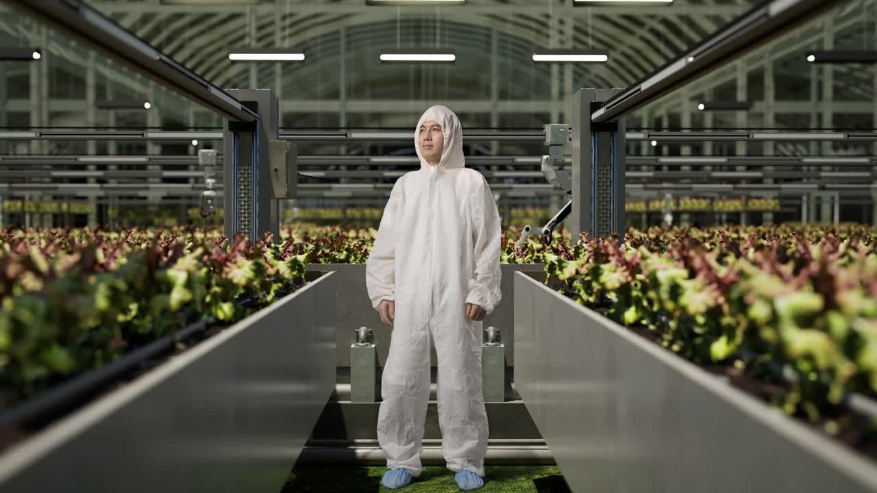 cuerpo lleno de satisfecho hombre asiático investigador mirando a su alrededor y sacudiendo la cabeza mientras estaba de pie en el invernadero con agricultores robóticos inteligentes