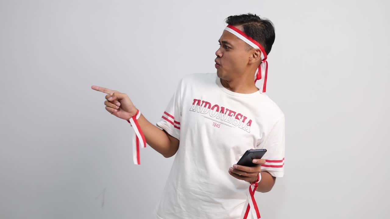 Seorang pria Asia yang bahagia memegang ponsel, dan menunjuk ruang kosong di sampingnya, terisolasi dengan latar belakang putih. Hari Kemerdekaan Indonesia